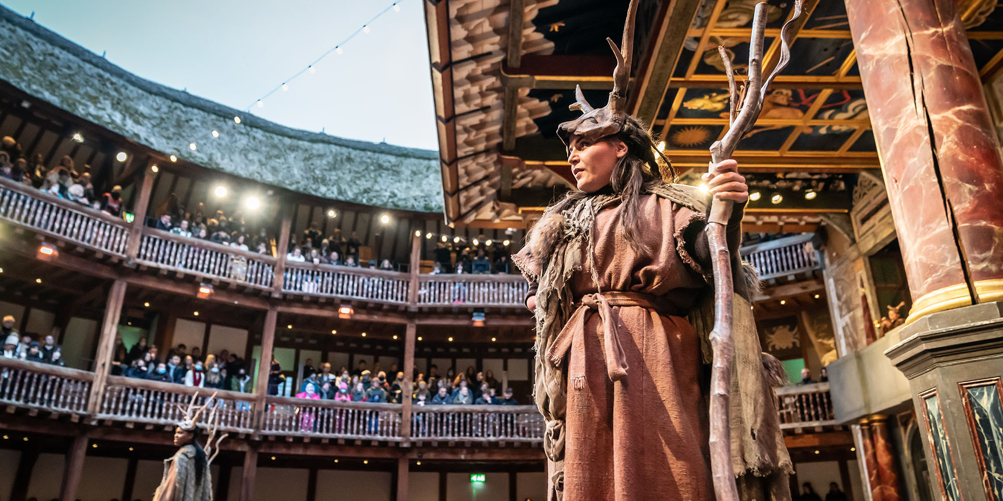 An actor wearing brown rags and a fur cloak, with an animal antler headpiece, holds a wooden staff on the Globe Theatre stage, looking up at the audience sat in the circular Galleries around them.