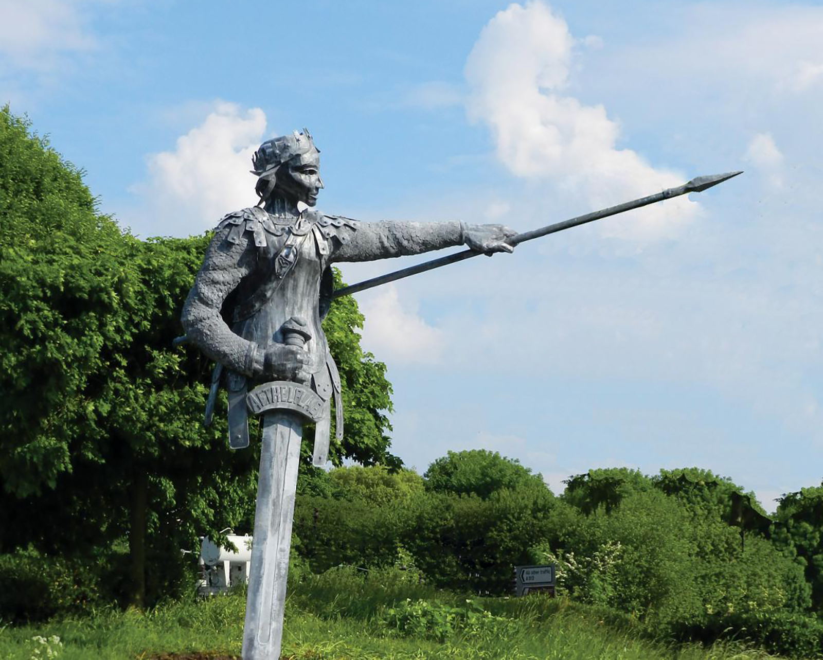 Aethelflaed A statue of Aethelflaed pointing a spear. The statue is in a green space with green trees and blue sky behind.